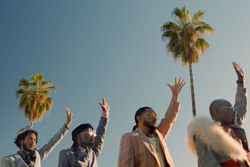 Four stylishly dressed men raise their left hands in front of two palm trees