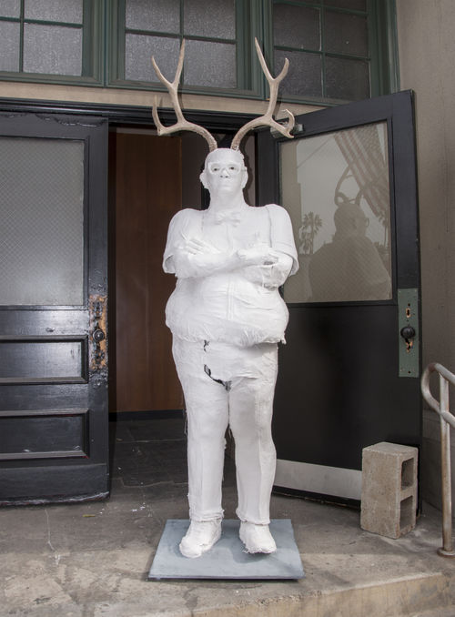 Life-sized plaster sculpture of a stern-looking man with deer antlers, standing arms crossed in front of an open door