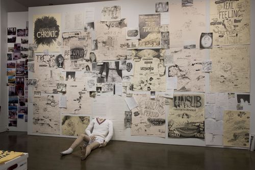 A gallery wall covered in drawings, with a headless mannequin sitting against it