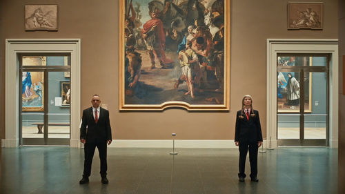 Two gallery attendants, a man and a woman, stand in front of a large painting