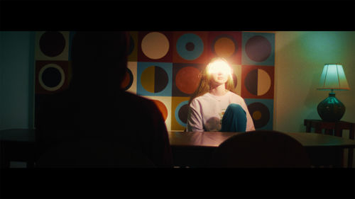 A young woman sits at a table, her eyes illuminated by a ray of sun through the window