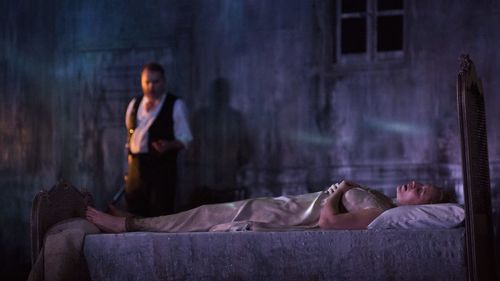 Man stands near woman sleeping in bed in dimly lit room. 