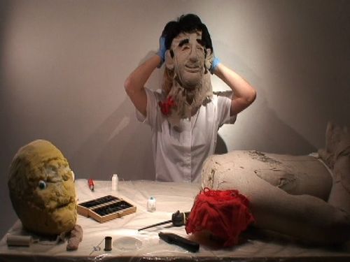 The artist removes a large, grotesque mask from her head while in front of her a decapitated dummy lies on a table