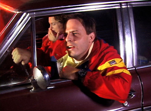 A man in a red jacket leans out the driver's side window of a car,