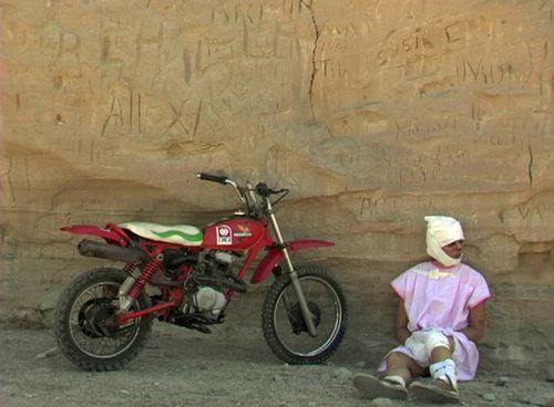 A woman in a pink hospital gown with her head bandaged sits against a rock next to a red dirtbike
