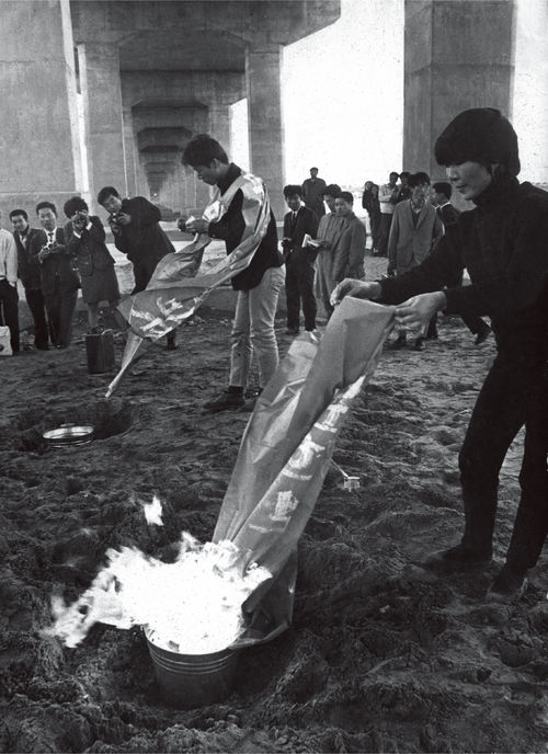 Image of people standing under a bridge, burning large pieces of paper in flaming buckets in the sand