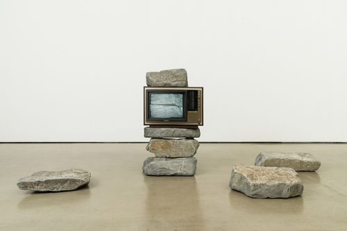 An old television set rests in the middle of a stack of large rocks, showing an image of two rocks on screen