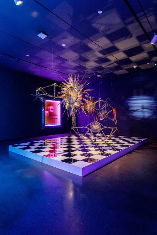 View of a gallery lit in green and pink, with a large, vibrant sculpture hanging from the ceiling