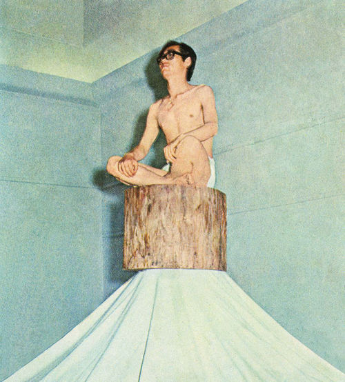 A Korean man stripped to his underwear sits cross-legged on a tree stump