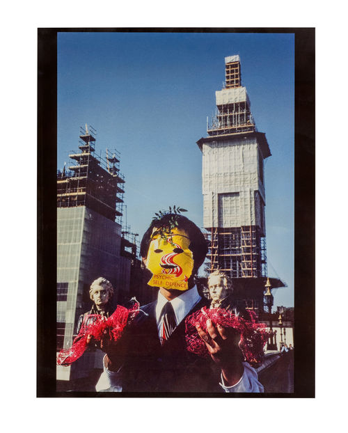 Photo of a man wearing a yellow mask reading "Psychic Self-Defence" carrying two miniature busts