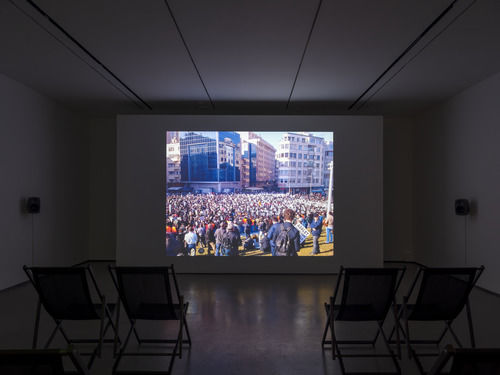 Installation view of single-channel video displaying a crowd of people between large buildings, a short row of black chairs made available for visitor seating