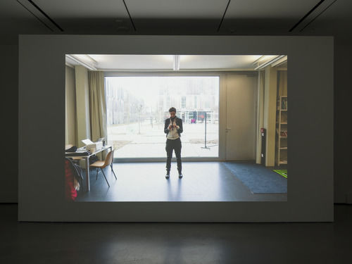 Installation view of single-channel video displaying a person standing in a room in front of a sunny window, a short row of black chairs made available for visitor seating