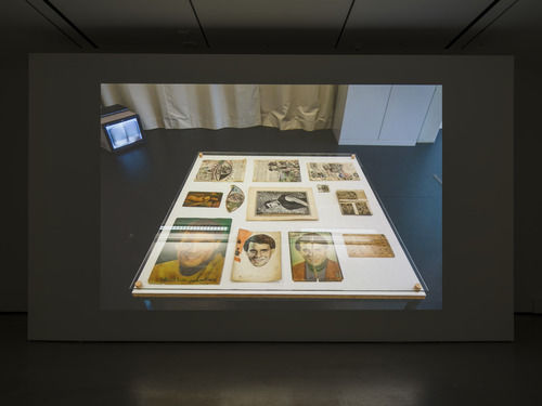 Installation view of single-channel video displaying a table of old photographs, a short row of black chairs made available for visitor seating