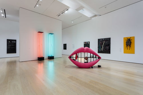 Installation image showing wall-mounted artworks and multimedia sculptures, including a mouth with pink lips and mirrors for teeth