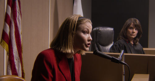 A blond, White woman sits on the witness stand, speaking into a microphone
