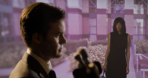 A man stands in front of microphones, while a woman looks at him from the background