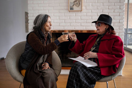 Cecilia Vicuña and Teresinha Soares sit in chairs and clink glasses