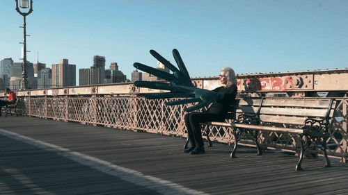 Artist Sylvia Palacios Whitman sitting on a bench on the Brooklyn Bridge, wearing an oversized hand
