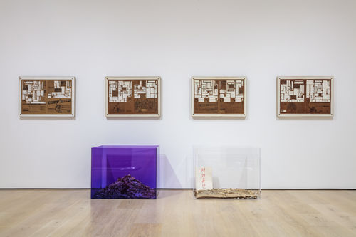 Framed newspaper sheets with words cut out hang on a wall, with two lucite boxes full of paper scraps sit in front