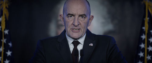 A man in a suit stands in front of two American flags, addressing the camera directly