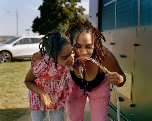 A Black woman and girl drink water from a hose