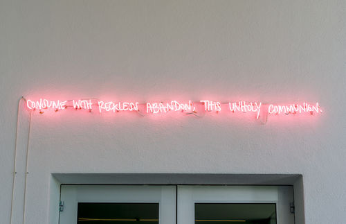 A pink neon sign, installed over a gallery door, which reads: Consume with reckless abandon, this unholy communion