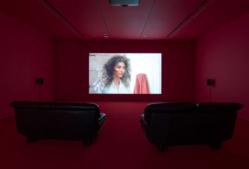 Single-channel video installation in a room with red walls and two leather couches