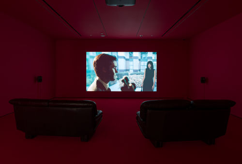Single-channel video installation in a room with red walls and two leather couches