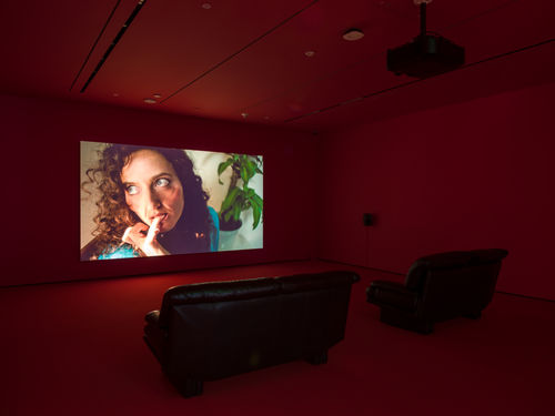 Single-channel video installation in a room with red walls and two leather couches
