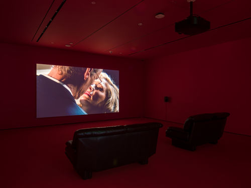 Single-channel video installation in a room with red walls and two leather couches