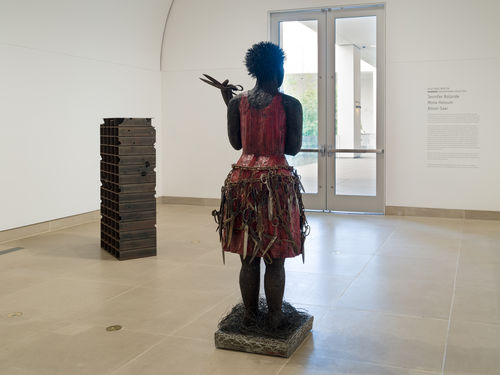 installation image showing free-standing sculptural works: one depicting female figure with scissors in her hand and arranged in a belt around her waist; the second made of rows of mild steel tubing