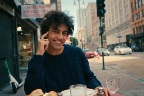 A figure in a black sweater sits at a cafe table on a sidewalk
