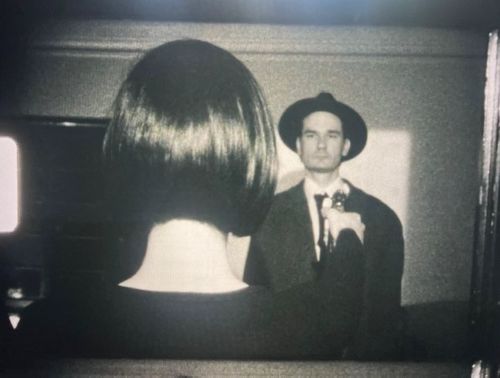 Black-and-white image of the back of a person's head with short black hair, pointing a metal object at another figure wearing a black suit and hat