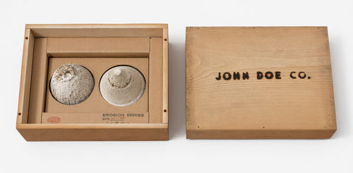 A simple wooden box: on the left, two small mounds of sand sit in the box, with a stamp reading "EROSION SERIES," on the right, the box top with the words "JOHN DOE CO." burned into it