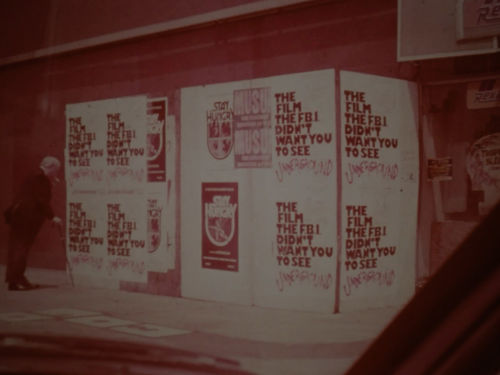 Pink toned still of a man approaching a wall papered over with posters reading "The film the FBI doesn't want you to see"