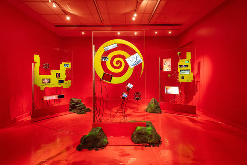 Installation image of a red room with sculptures made of transparent glass, yellow shapes, and electronic components