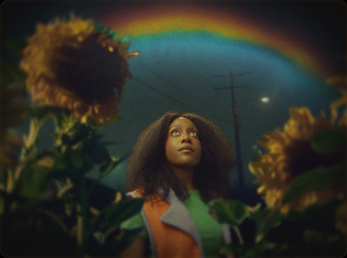 A Black woman, framed by sunflowers, looks up at a rainbow in the night sky