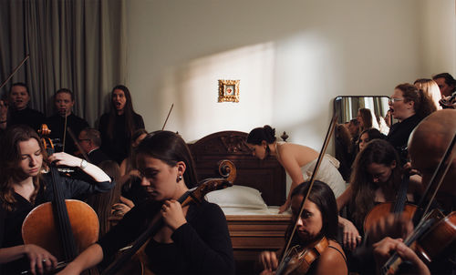 A woman leans over a bed, surrounded in the room by a symphony orchestra