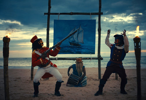 Three performers in punch-and-judy make up on a beach, one pointing a musket at another