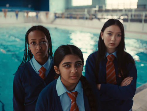 Three girls in school uniforms look at the camera, with a pool behind them