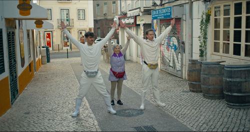 Two young men in all white and an older woman in purple stand in a city street with their arms stretched above their heads