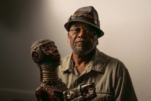 The artists Akinsanya Kambon sits next to his sculpture of a long-necked figure