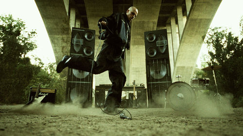 A man stands in front of amps and instruments under a highway overpass, holding a microphone by the cord