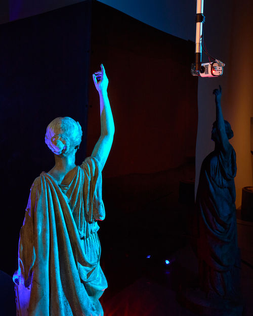 A bronze sculpture of a female figure lifting her right arm, relfected in a black wall, with a surveillance camera overhead