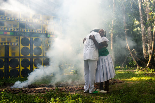 A man and woman embrace in front of a wall of yellow speakers, surrounded by smoke