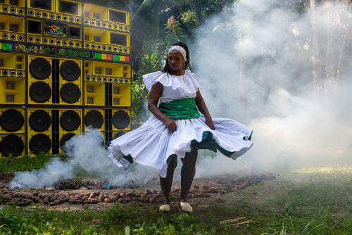 A woman in a white dress dances in front of a wall of yellow speakers, surrounded by smoke
