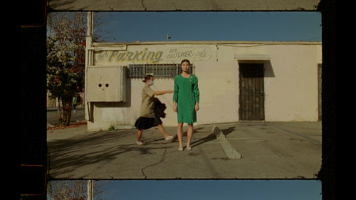 Two people stand in an empty parking lot, one faces the viewer; another walks behind them heading towards their right