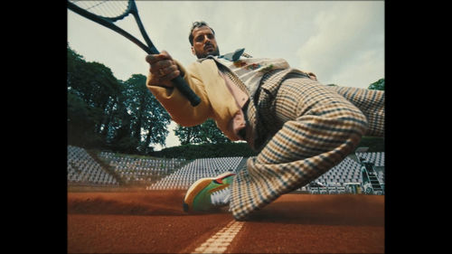 A tennis player dressed in street clothes looks directly into the camera as they bend over towards the court floor
