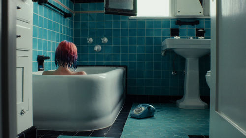 A figure sits in a bathtub with wet, died pink and blue hair, facing away from the camera