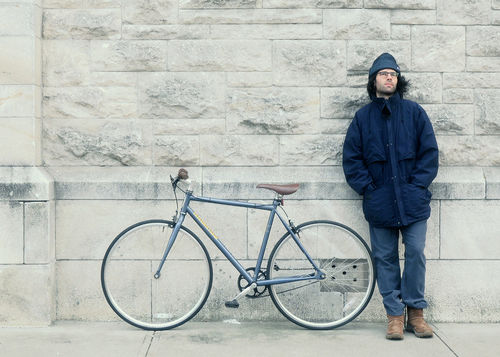 A figure dressed in a heavy coat leans against a wall, standing next to a bike